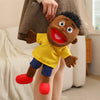 A soft plush full-body hand puppet of a Black child with dark curly hair wearing a yellow shirt and blue shorts.