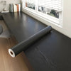 A roll of self-adhesive wood grain wallpaper lying on a dark wooden desk near a window.