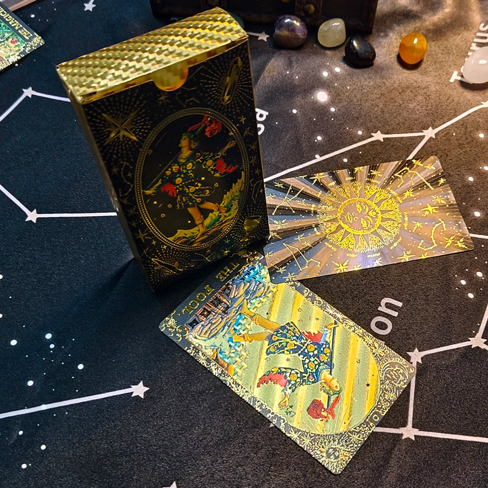 A gold foil tarot card deck and guidebook are displayed on a dark surface surrounded by polished stones.