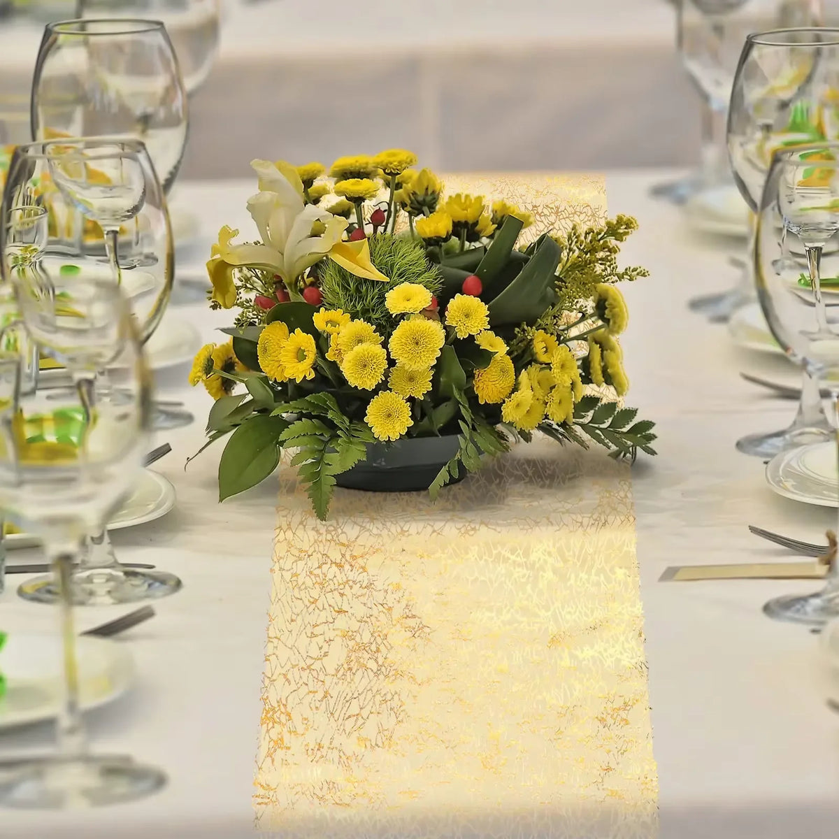 A metallic gold glitter table runner is placed on a white tablecloth, featuring a yellow floral arrangement.