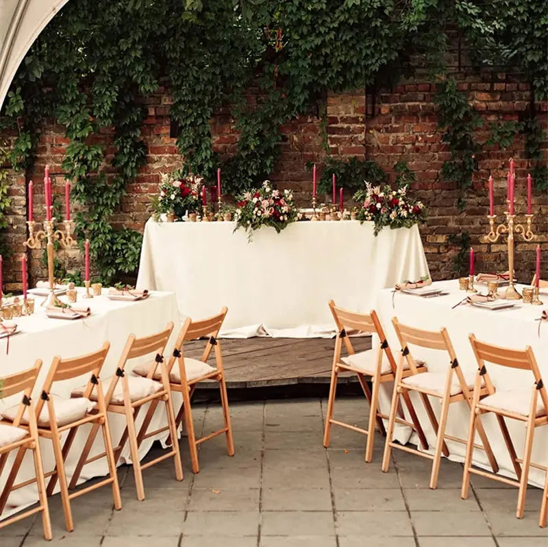 A long white satin tablecloth is set up outdoors against a brick wall with greenery and candles, ready for a wedding or forma