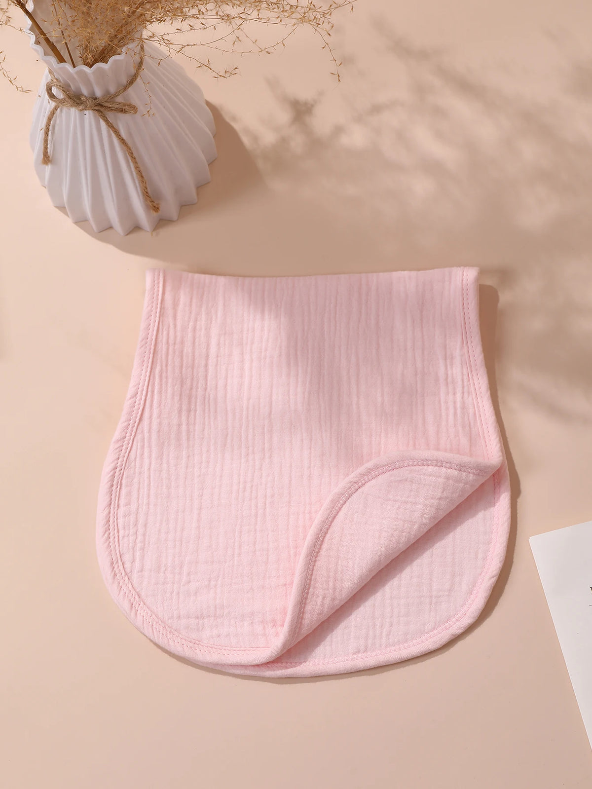 A pink, ribbed baby burp cloth is folded on a light beige surface next to a vase of dried grasses.