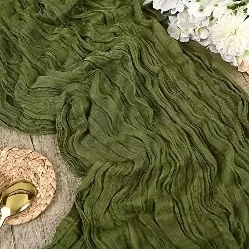 A close-up of a moss green cotton gauze table runner draped in soft, wavy folds with white flowers visible in the background.