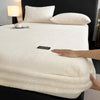 A white plush mattress cover is displayed on a bed, with a hand touching the fabric.