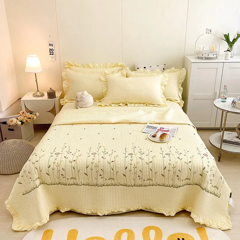 A yellow embroidered floral quilted bedspread set displayed on a bed with white linens and light wood flooring.