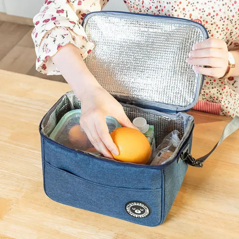 A hand is opening a blue insulated lunch bag with a silver foil lid, revealing an orange fruit inside.