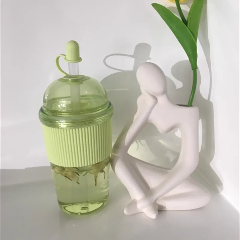 Helles grünes Glas-Tumbler mit Strohhalmdeckel neben einer weißen, stilisierten Figur und einer grünen Pflanze.