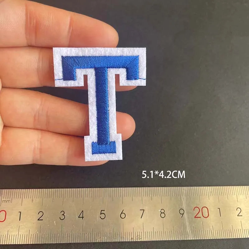A hand holds a blue embroidered letter T patch, measuring 5.1 x 4.2 cm against a ruler.