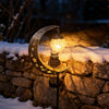 A solar moon-shaped garden light featuring an owl silhouette and a glowing glass ball, set on a stone wall covered in snow.