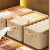 A hand is opening a beige fabric storage box filled with neatly folded fabric items for drawer and closet organization.