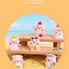 Pink resin pig figurines in various poses displayed on a wooden surface with yellow sand and small stones.