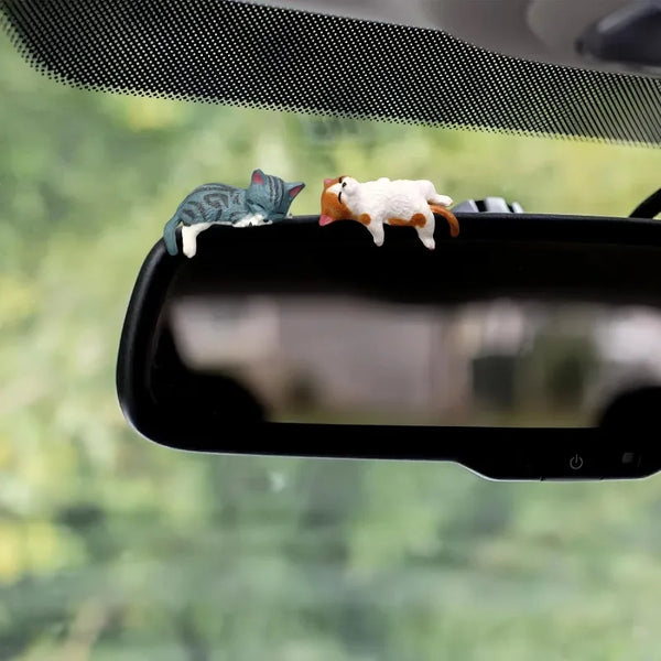 A set of two resin cartoon cat ornaments, one gray and one white with orange accents, displayed on a car dashboard.