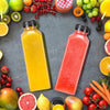 A flat lay of various fresh fruit juices and whole fruits in colorful plastic bottles on a gray surface.
