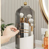 A clear glass rotating brush holder filled with makeup brushes and a gold dustproof lid on a white surface.