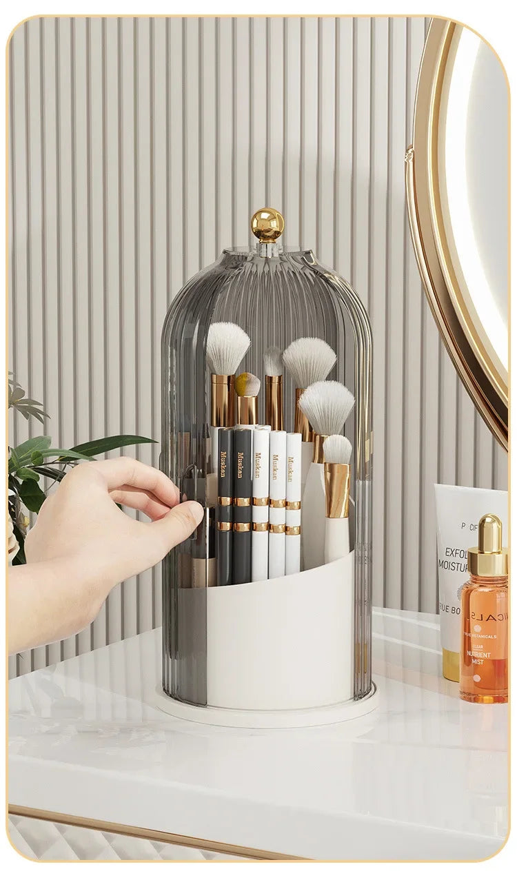 A clear glass rotating brush holder filled with makeup brushes and a gold dustproof lid on a white surface.