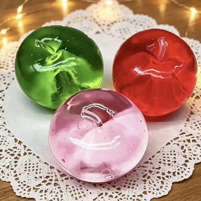 Three fruit-shaped slow-rising squeeze toys in green, red, and pink resting on white lace