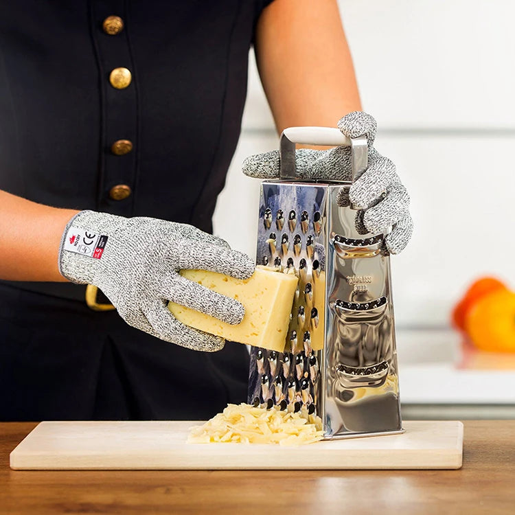 A hand wearing silver cut-resistant work gloves is holding a block of cheese over a wooden cutting board.