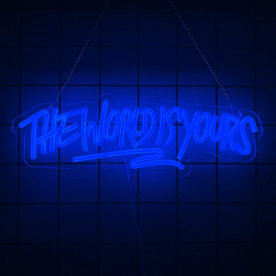 A glowing blue neon sign shaped like the words The World Is Yours is suspended against a dark grid background.