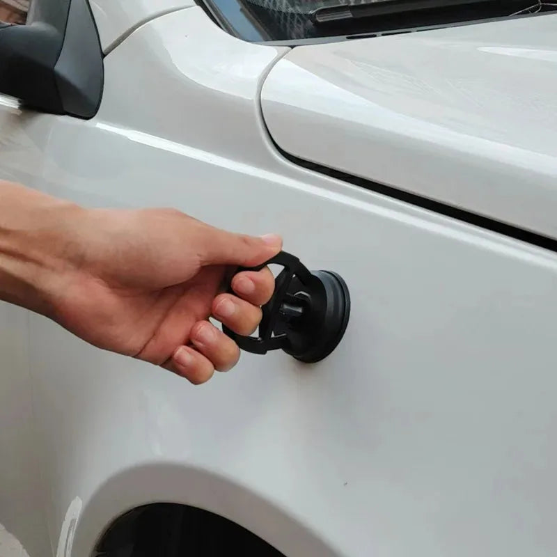 A hand is operating a black handle on the side of a white car panel, demonstrating a car dent puller kit.