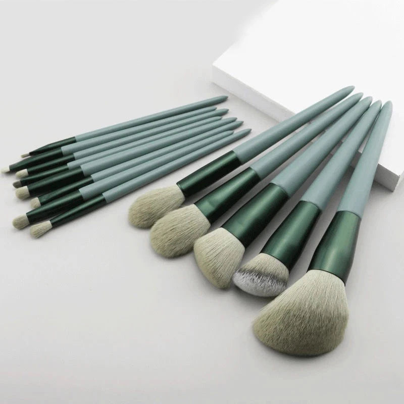 A set of thirteen makeup brushes with green and gray handles arranged on a white surface.