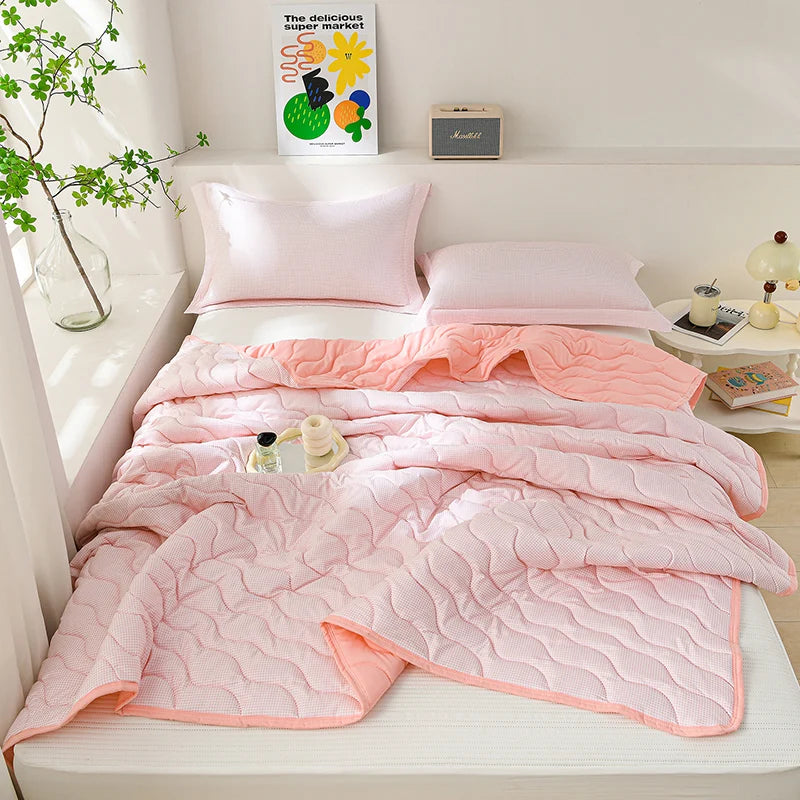 A pink quilted blanket is draped over a white bed, next to a plant and some decorative items.