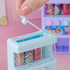 A hand is inserting a white plastic piece into an interactive vending machine toy that dispenses play coins and drinks.