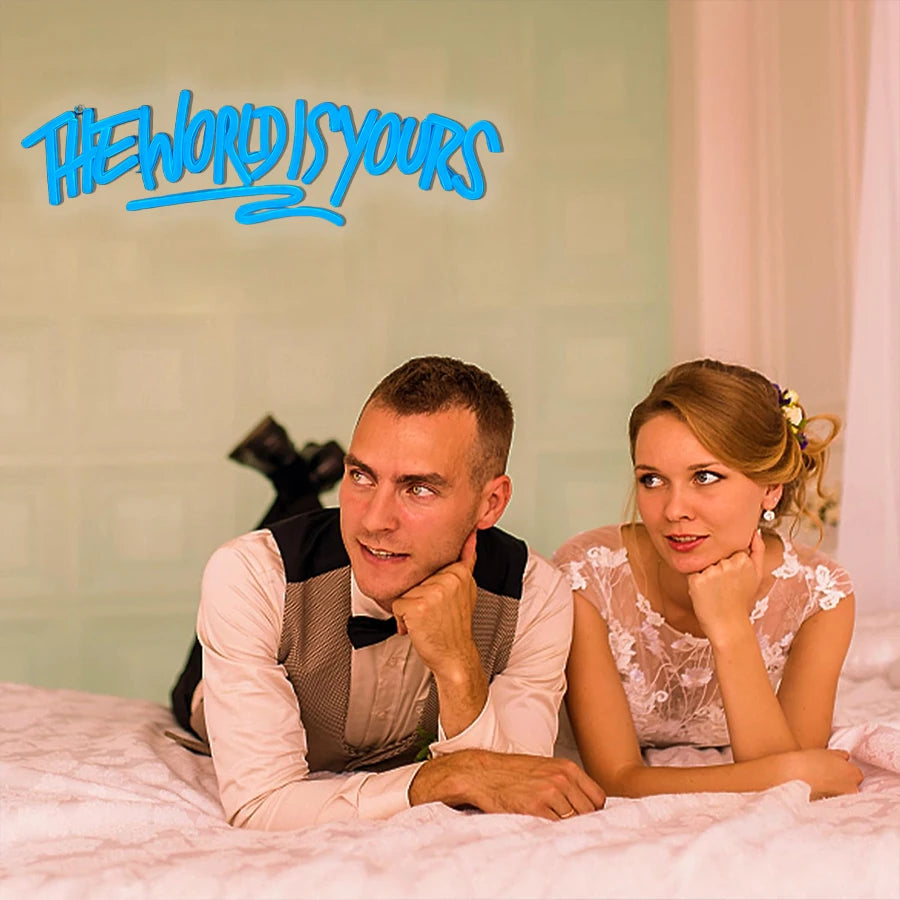 A man and a woman looking at the camera while lying on a white surface with a blue neon sign in the background.