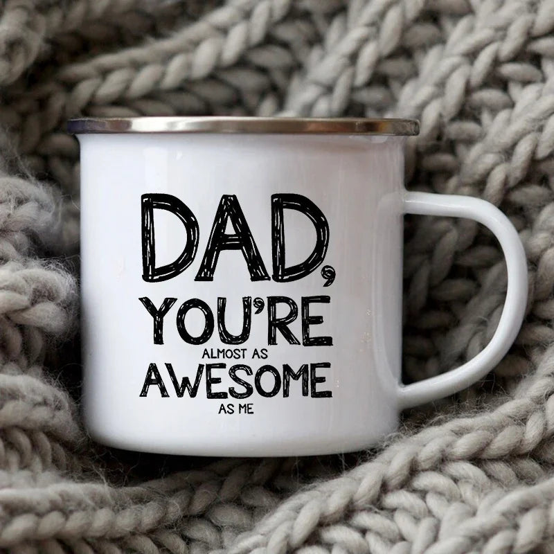 Tasse à café en émail blanc avec l'inscription : 'Papa, tu es presque aussi génial que moi', posée sur une couverture tricoté