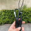 A hand holds a black bungee tether with a carabiner hook and adjustable straps against a green hedge background.