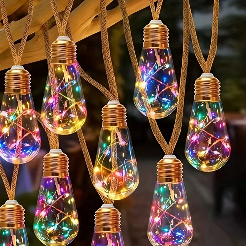 String lights with glowing filaments are suspended by hemp rope against a blurred outdoor background.