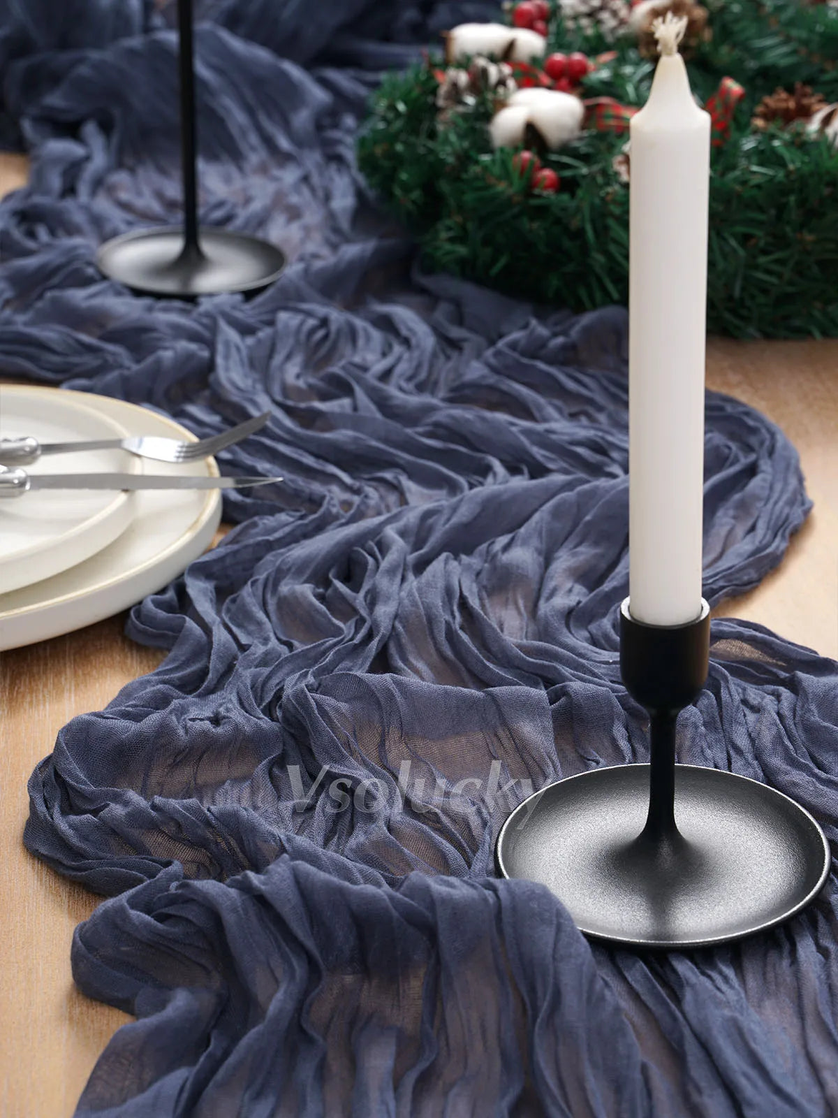 A dark blue crinkle gauze cheesecloth table runner draped over a wooden surface with a white candle and silver cutlery.