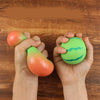 Hände halten zwei wassermelonenfarbene Stressball-Spielzeuge auf einer Holzoberfläche.
