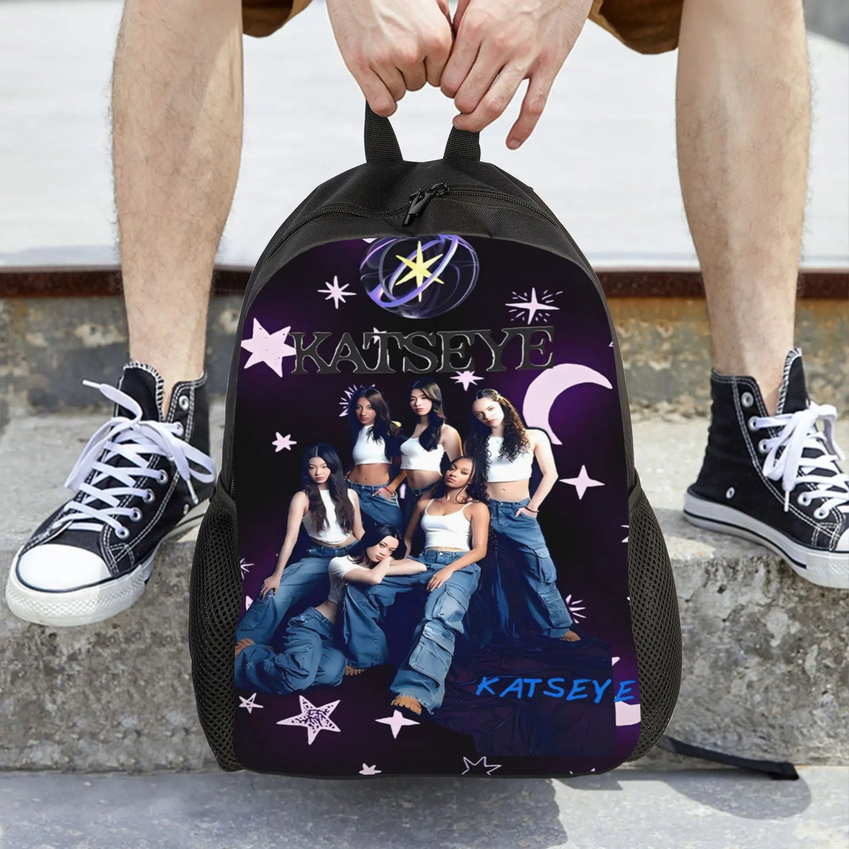 A black canvas backpack featuring the name KATSEYE in purple and white stars on the front, held by a person.