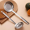 A stainless steel fine mesh skimmer spoon resting on a wooden board next to peppercorns and an orange.