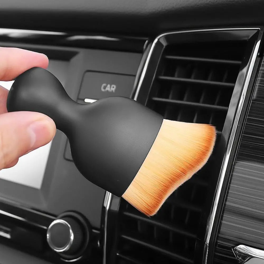 A hand holds a black ultra-soft curved detailing brush with a light wood-colored ferrule, positioned near a car vent grille.