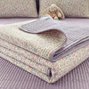A stack of floral quilted bedspreads in lavender and green patterns with dust-proof covers resting on a purple surface.