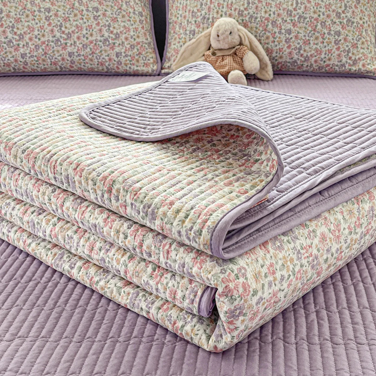 A stack of floral quilted bedspreads in lavender and green patterns with dust-proof covers resting on a purple surface.