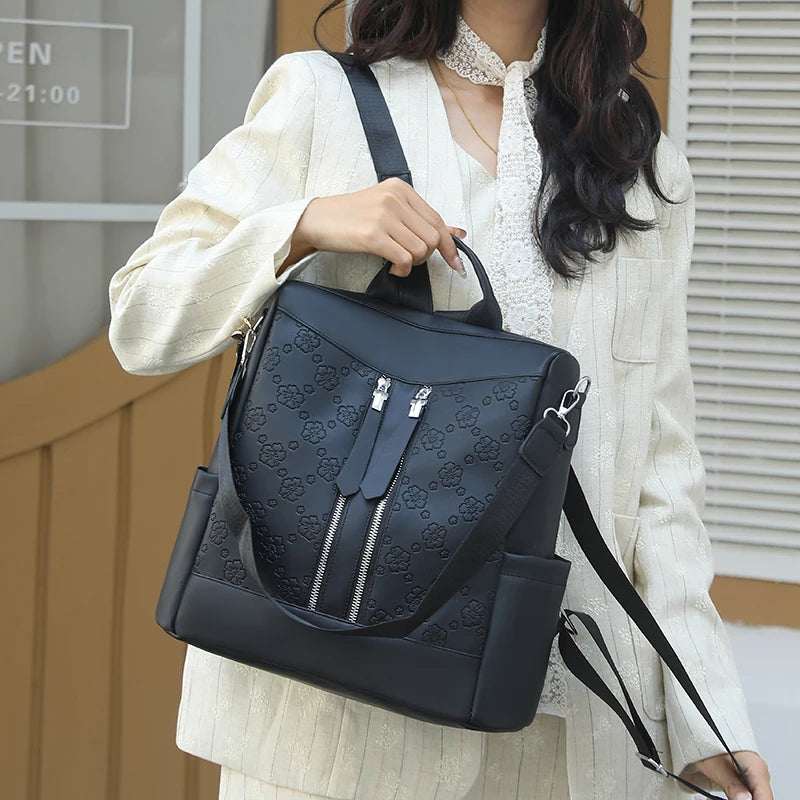A woman is holding a black anti-theft leather backpack with silver zippers and a patterned design.