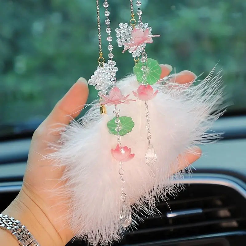 A hand holds a pink lotus car pendant featuring clear crystals and green leaves on a white feather background.