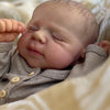 A close-up photo of a newborn baby doll with closed eyes resting on a soft, patterned fabric.