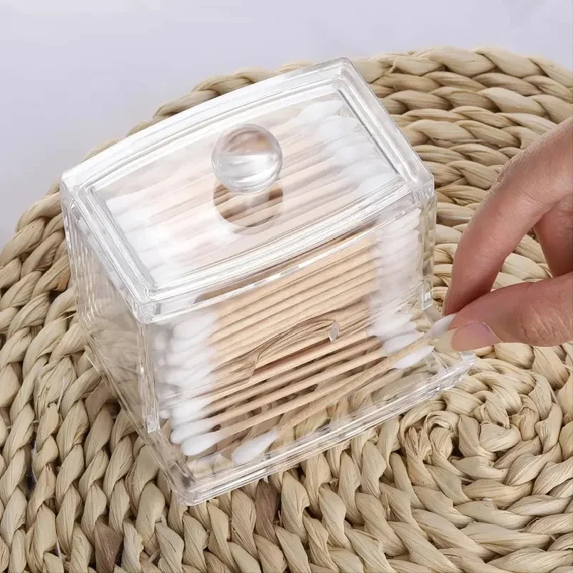 Clear plastic storage dispenser for cotton swabs and pads with a wooden lid resting on a woven mat.