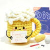 A yellow crocheted animal mug with pink accents sits next to yarn and a crochet kit.