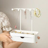 A white elegant jewelry display rack with a drawer and wooden base is shown hanging on a wall.