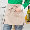 A beige half apron with a tie closure and pockets is shown being worn over a green shirt.