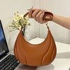 A hand holds a brown textured leather crossbody bag with a gold chain strap resting on a white desk.