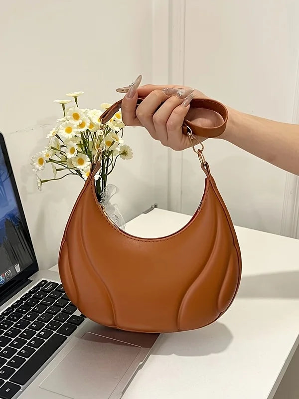A hand holds a brown textured leather crossbody bag with a gold chain strap resting on a white desk.
