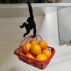 A black monkey-shaped wooden wall hook is hanging above a red basket filled with oranges.