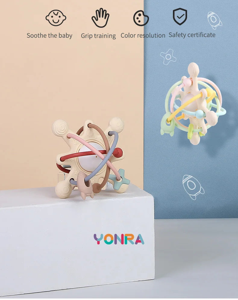 A silicone sensory rattle ball with various colored rings is displayed on a white box against a blue background.