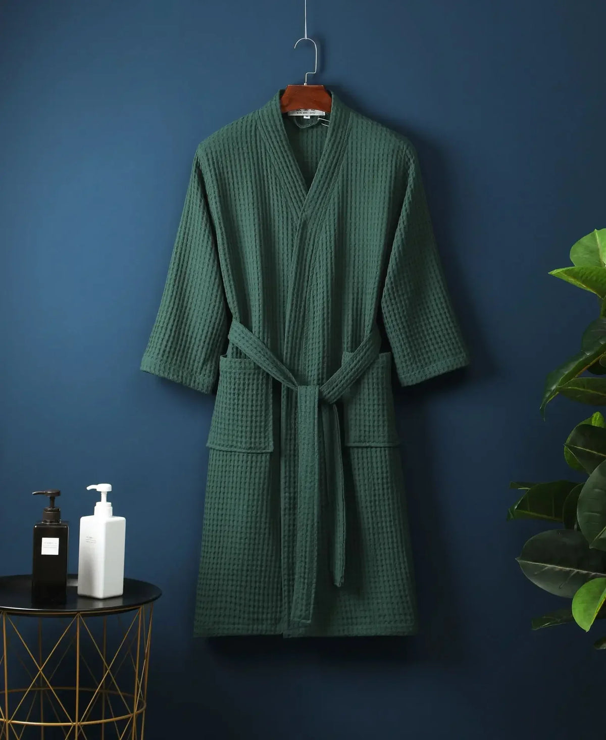 A dark green lightweight cotton waffle robe hanging on a wall against a dark blue background.
