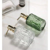 A clear glass soap dispenser with a gold pump mechanism and a green liquid refill bottle.